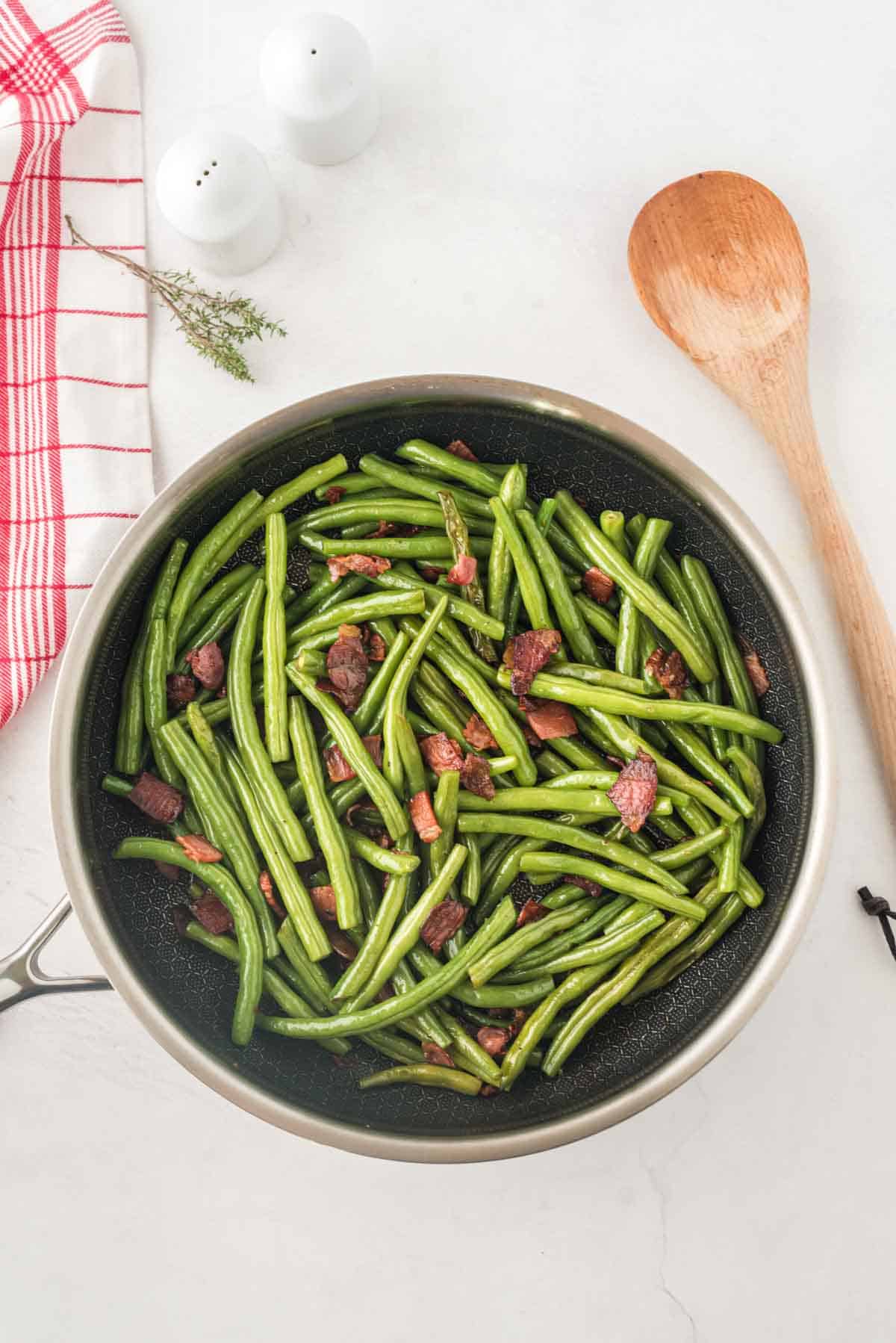 Sauteed Green Beans with Bacon in a skillet with a wooden spoon next to it.