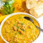 A bowl of split pea and ham soup with a spoon, with hunks of bread in the background.