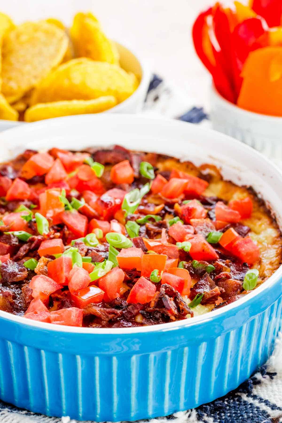 A baked dip topped with bacon and tomatoes with bowls of tortilla chips and pepepr slices.
