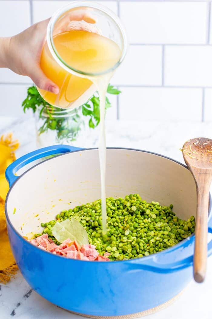 A hand pouring stock from a jar into a pot with split peas and ham.