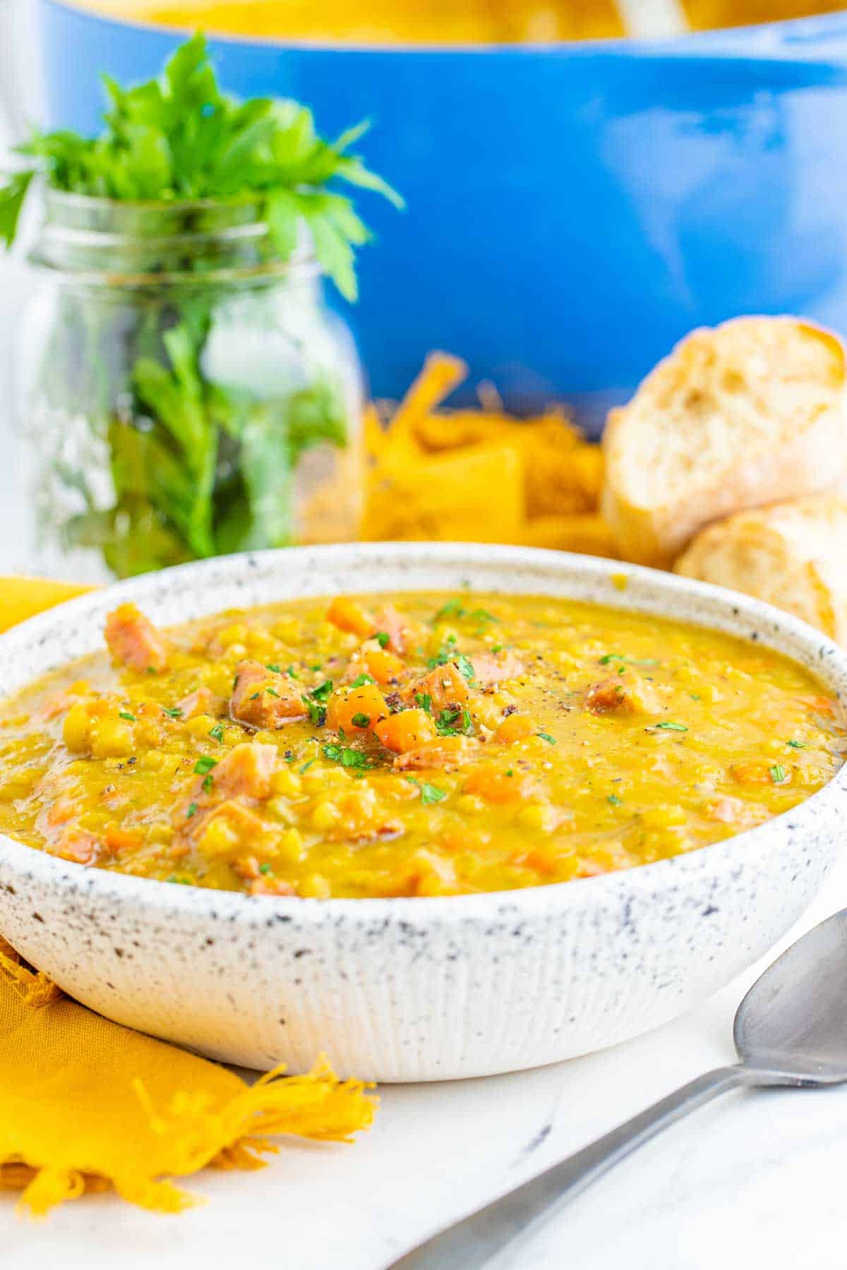 A bowl of split pea soup with ham with a blue pot and a jar of fresh herbs in the background.