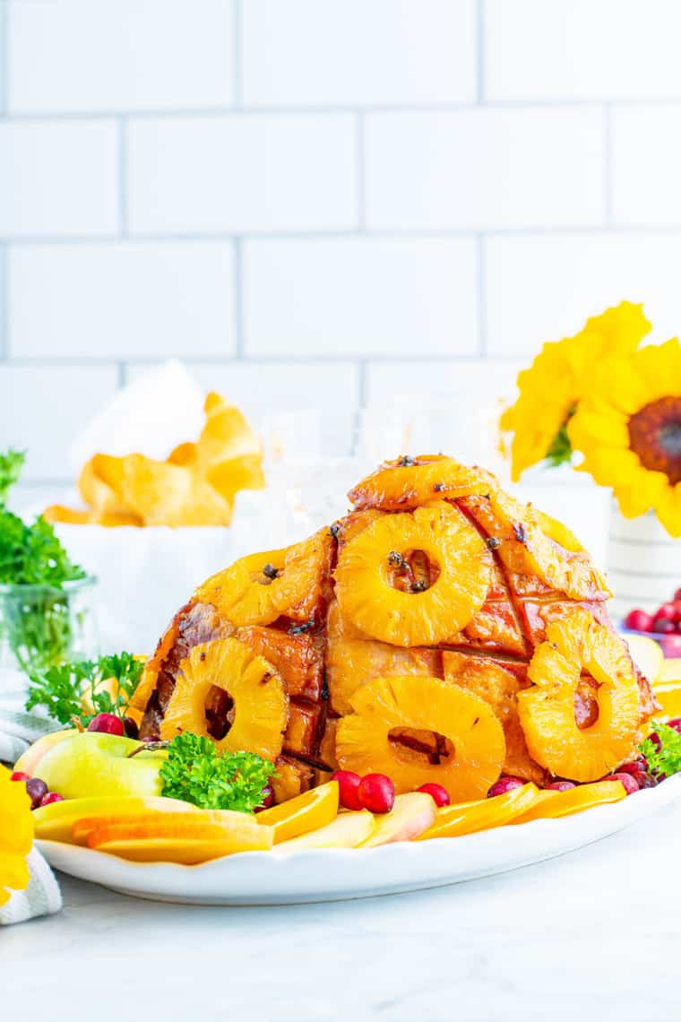 Baked ham with pineapple slices on the outside on a serving platter garnished with apples, oranges, and cranberries