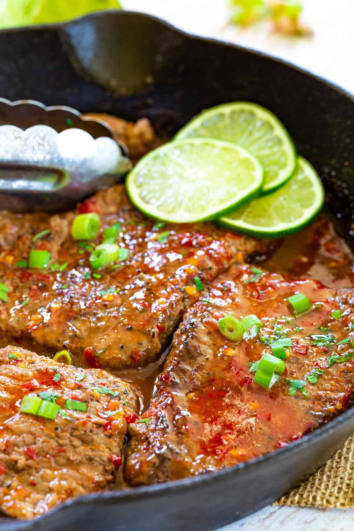 Chili lime cube steaks in a cast iron skillet with tongs and lime slices