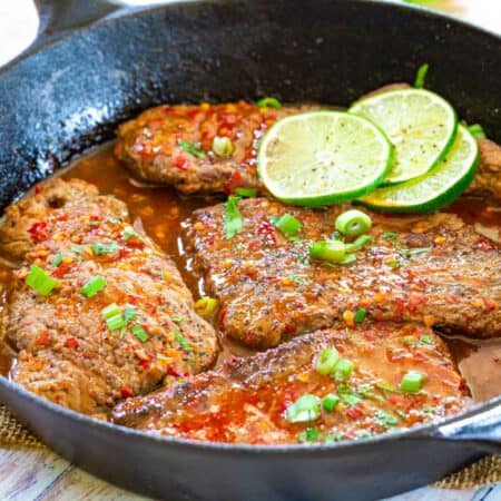 Cast iron pan with four cube steaks with a red-colored chili sauce and lime slices