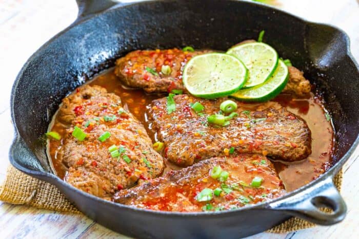 Cast iron pan with four cube steaks with a red-colored chili sauce and lime slices