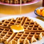 Pouring syrup on a waffle with butter with a stack of banana waffles on a platter in the background.