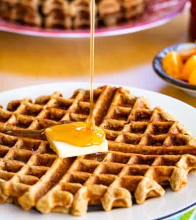 Pouring syrup on a waffle with butter with a stack of banana waffles on a platter in the background.