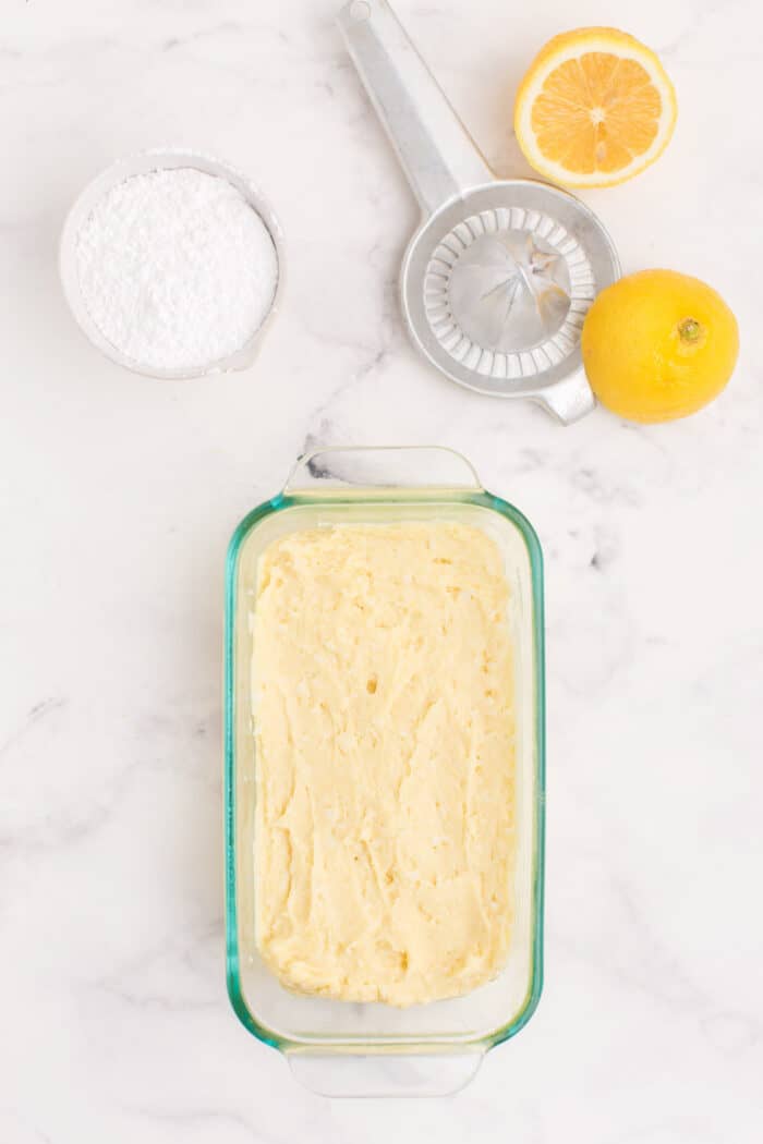 Unbaked batter in a loaf pan with the bowl of powdered sugar, lemon halves, and juicer on the side.