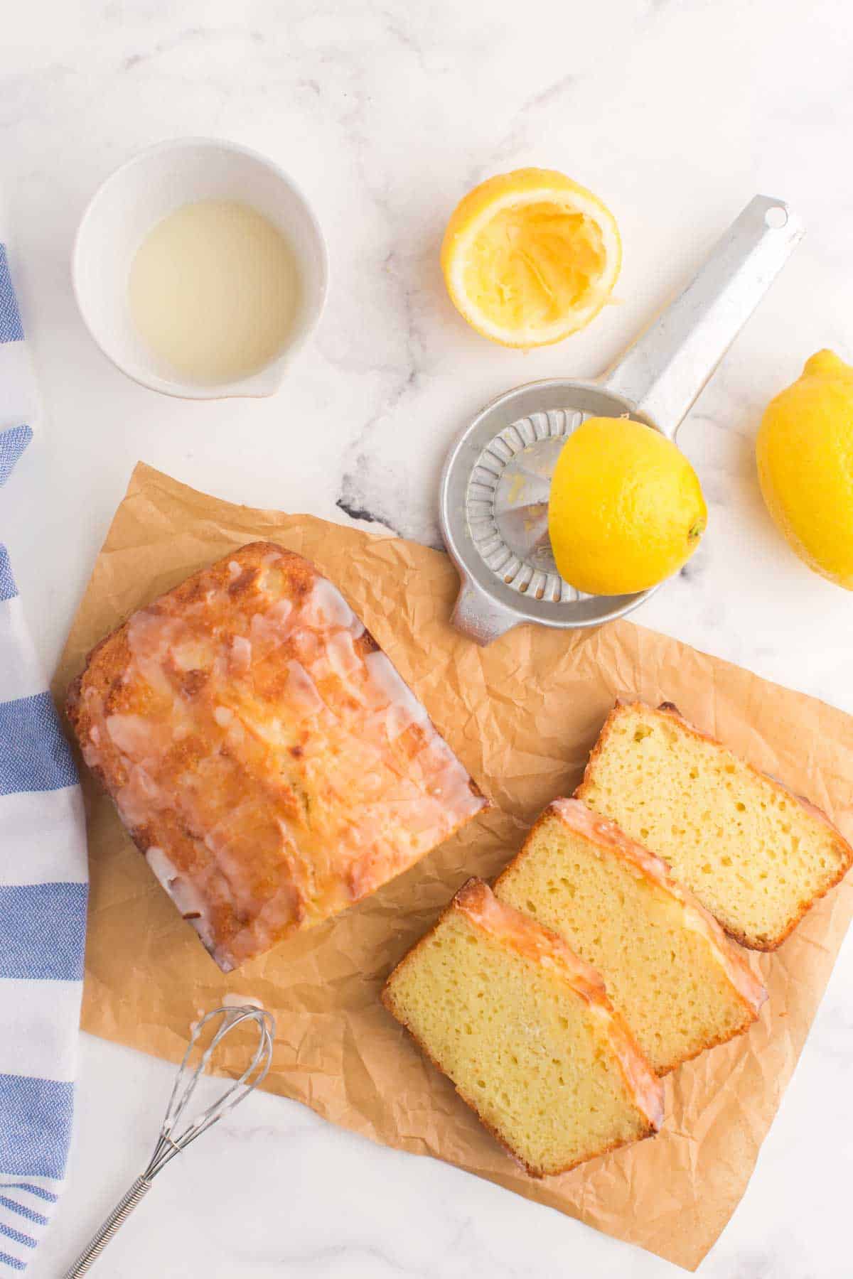 Half of the lemon pound cake on a piece of parchment alongside three slices and a whisk dripping with glaze.