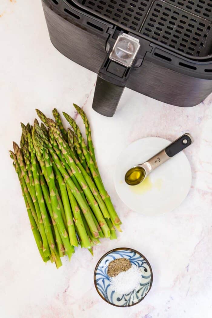 An air fryer, raw asparagus, a teaspoon of olive oil, and a small bowl of salt and pepper on a marble tabletop