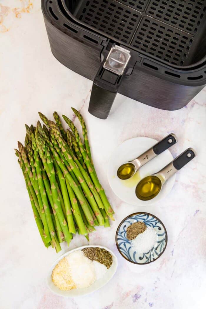 An air fryer, raw asparagus, a teaspoon and a tablespoon of olive oil, a small bowl of salt and pepper, and a ramekin with salt, pepper, cornmeal, parmesan cheese, and dried herbs on a marble tabletop