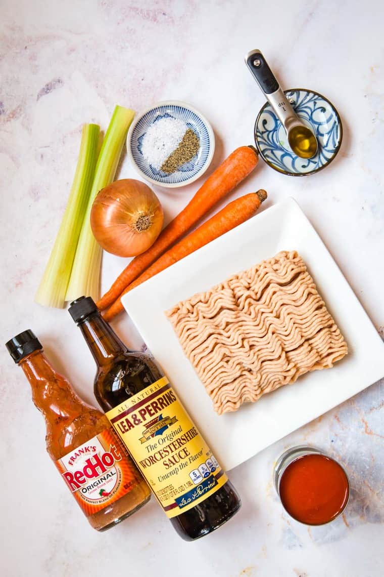 Ground chicken on a plate surrounded by bottles of hot sauce and worcestershire sauce, a can of tomato sauce, a teaspoon of oil, salt and pepper, and carrots, celery, and an onion