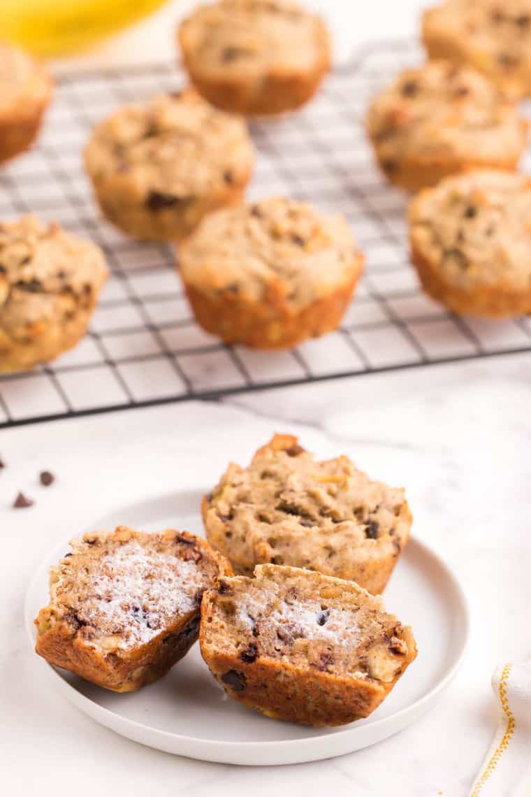 one buttered and one whole gluten free banana muffin on a plate with more muffins on a cooling rack behind it