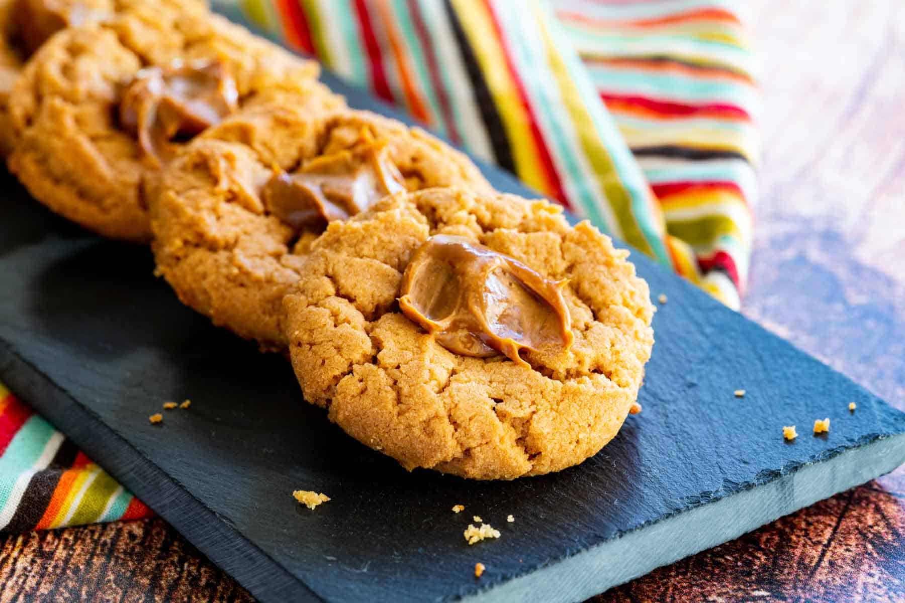 A slate platter with a lined up stack of dulce de leche peanut butter thumbprint cookies.