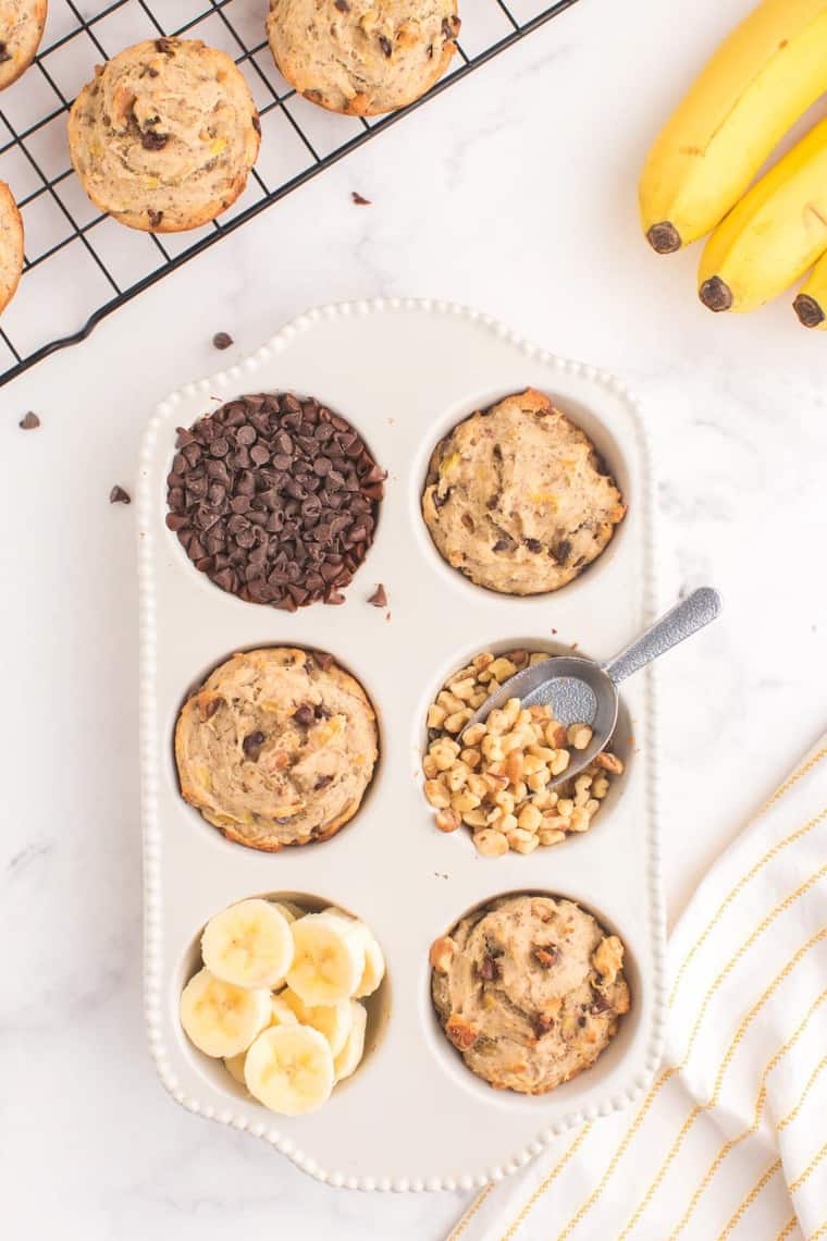 three baked muffins in a ceramic muffin pan with chocolate chips, walnuts, and bananas in the other cups