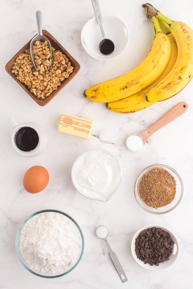 gluten free flour, salt, baking soda, bananas, vanilla, maple syrup, an egg, walnuts, ground flaxseed, butter, and mini chocolate chips in various bowls and spoons on a marble tabletop