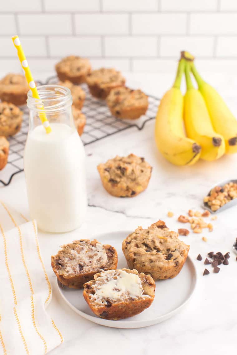 two banana muffins on a plate, one cut open with butter on a table surrounds by more muffins, bananas, and a bottle of milk