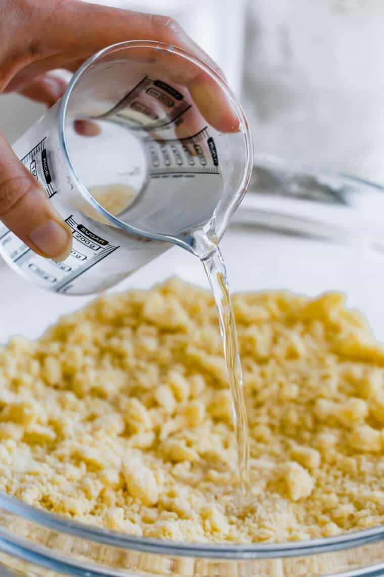 A hand pouring water from a measuring cup into a bowl of gluten-free pie dough.