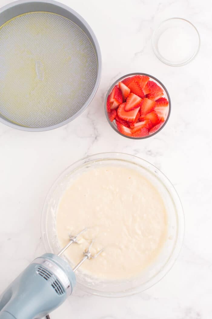 hand mixer in a bowl of cake batter next to a cake pan and bowl of strawberries