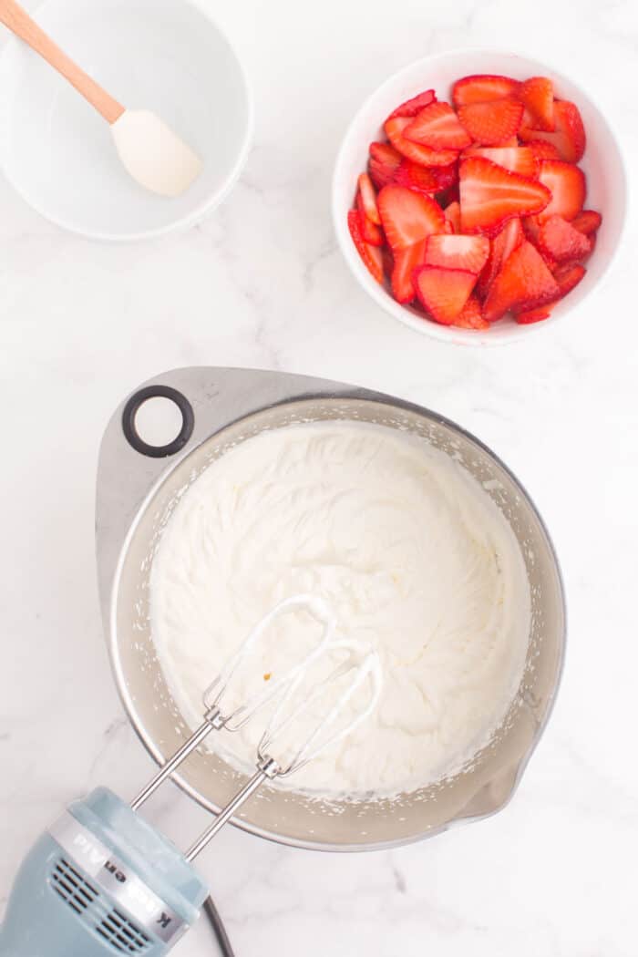 beaters in a bowl of fresh whipped cream and a bowl of sliced strawberries