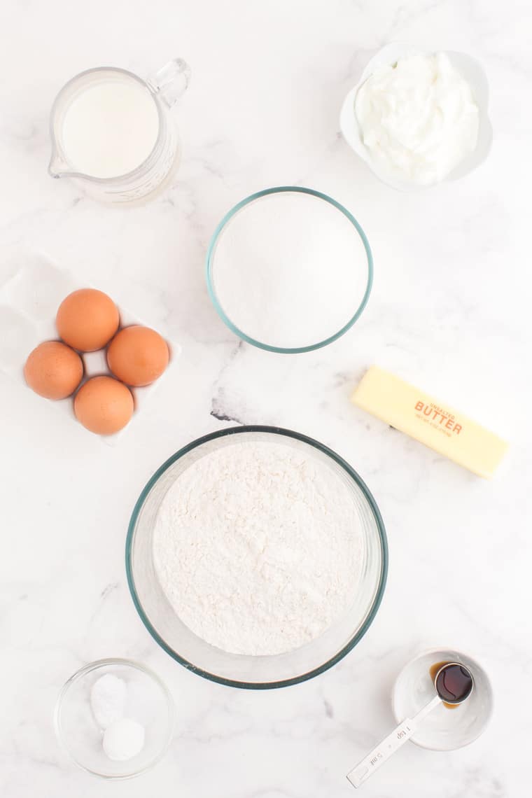 Gluten free flour, baking soda, baking powder, vanilla extract, eggs, butter, sugar, milk, and Greek yogurt in bowls and containers on a table