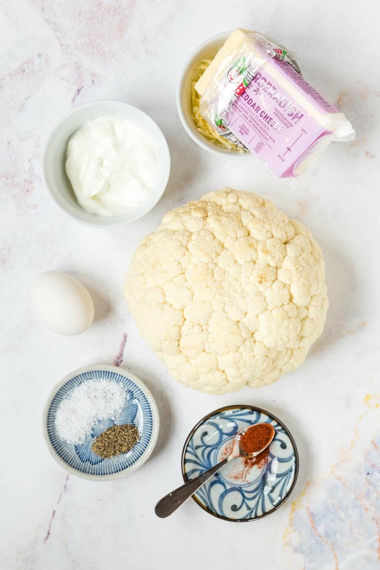 A head if cauliflower, an egg, and bowls with horseradish cheddar cheese, Greek yogurt, salt, pepper, and paprika on a marble tabletop