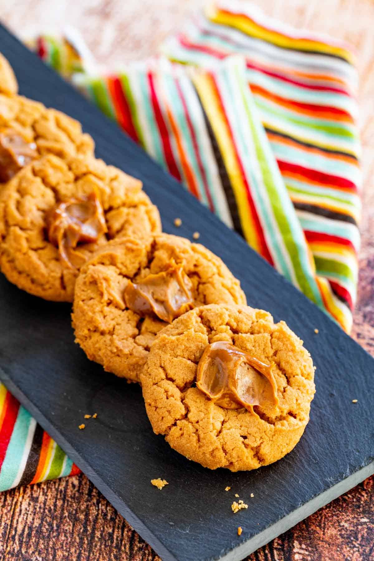 A line of peanut butter thumbprint cookies with a dollop of dulce de leche in the center of each on a slate platter.