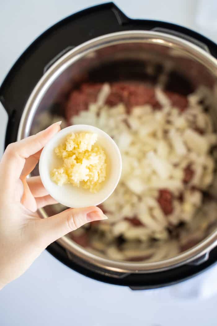 A Small Dish of Minced Garlic Being Held Over a Pressure Cooker Containing Beef and Cheese