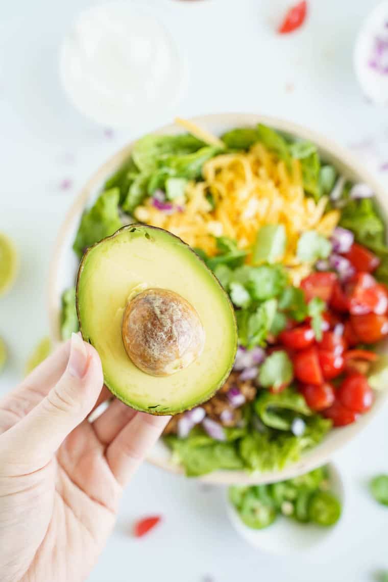 An Avocado Being Held Over a Colorful Keto Taco Bowl