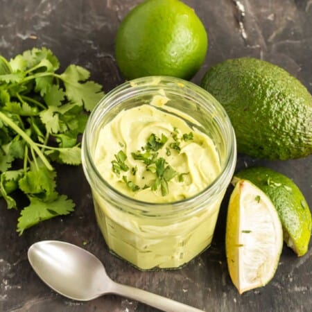 jar of avocado crema on a slate counter surrounded by an avocado, limes and a bunch of cilantro