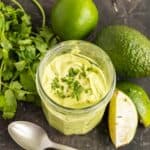 jar of avocado cream surrounded by an avocado, limes and a bunch of cilantro