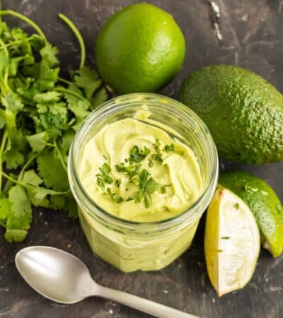 jar of avocado cream surrounded by an avocado, limes and a bunch of cilantro
