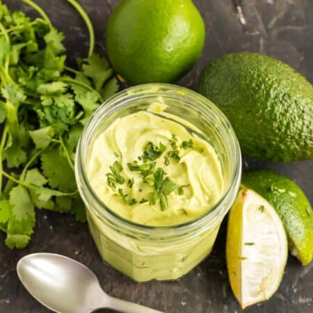 jar of avocado cream surrounded by an avocado, limes and a bunch of cilantro