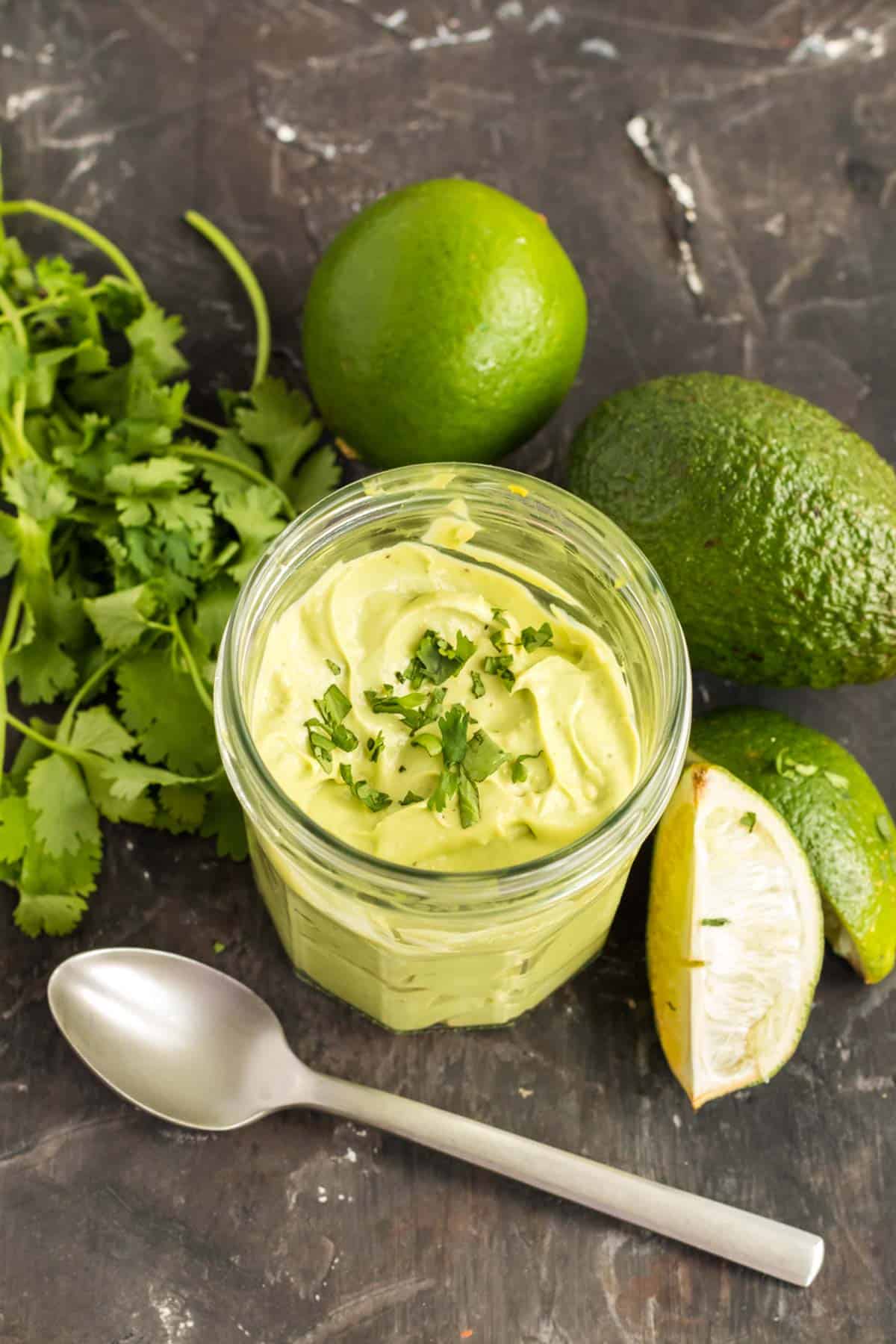 jar of avocado cream surrounded by an avocado, limes and a bunch of cilantro