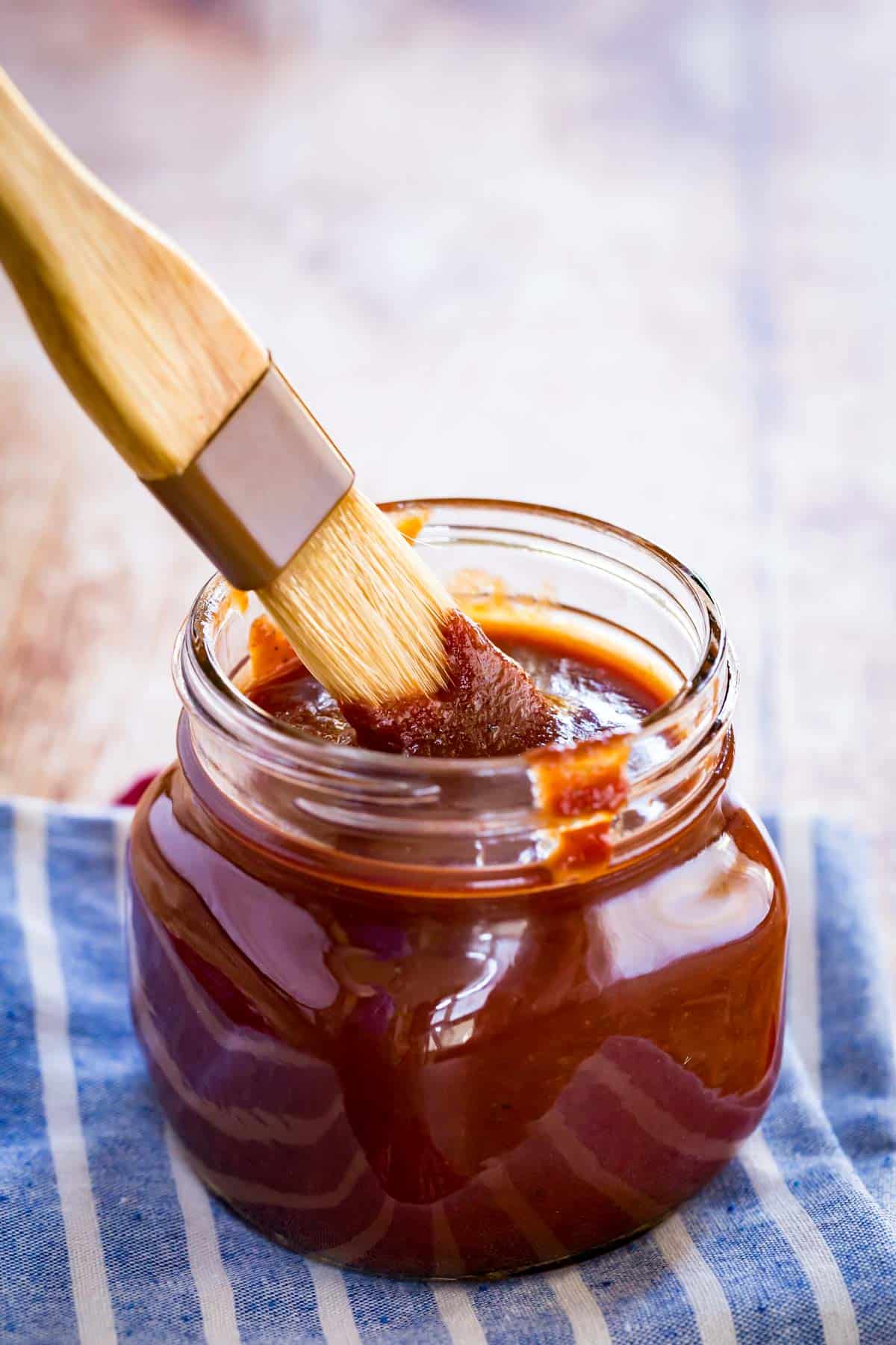 brush dunked into a glass jar of bourbon bbq on a white and blue striped cloth napkin