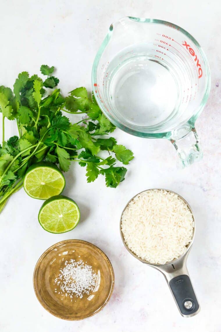 measuring cup of rice, measuring cup of water, bowl of salt, bunch of cilantro, and a lime cut in half on a marble tabletop