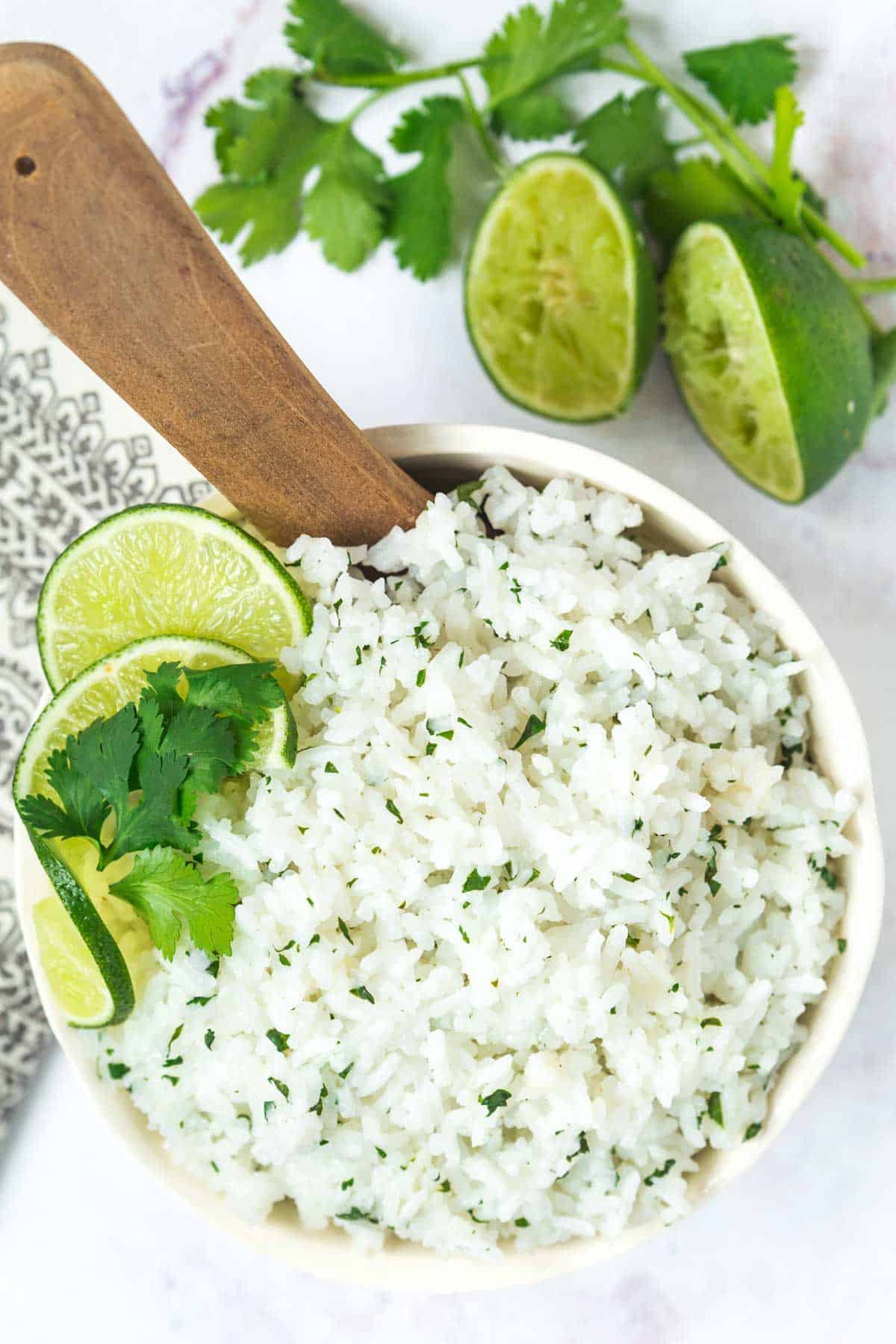 looking down at a bowl of cilantro lime rice with lime slices and cilantro leaves on the edge and a wooden spoon