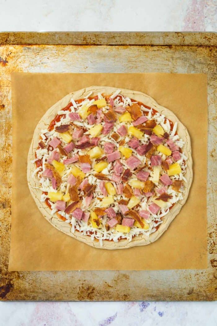 unbaked hawaiian pizza with ham, pineapple, and bacon on a baking sheet