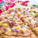 A spatula picking up a slice of ham and pineapple pizza with text overlay that says "Gluten Free Hawaiian Pizza".