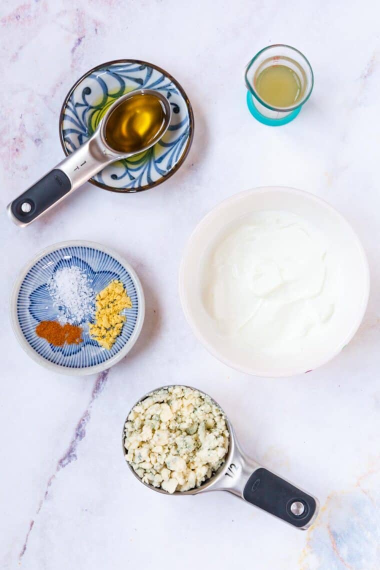 a bowl of plain Greek yogurt surrounded by a measuring cup of blue cheese, a small bowl with salt, dry mustard, and cayenne pepper, a tablespoon of olive oil, and a small beaker of vinegar