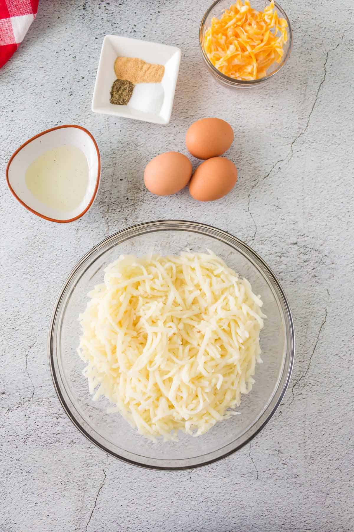 bowl of shredded potatoes, three eggs, and bowls of oil, shredded cheese, and seasonings on a tabletop