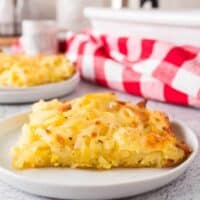 piece of the baked casserole on a white plate with a red and white checked napkin on a countertop with utensils in the background