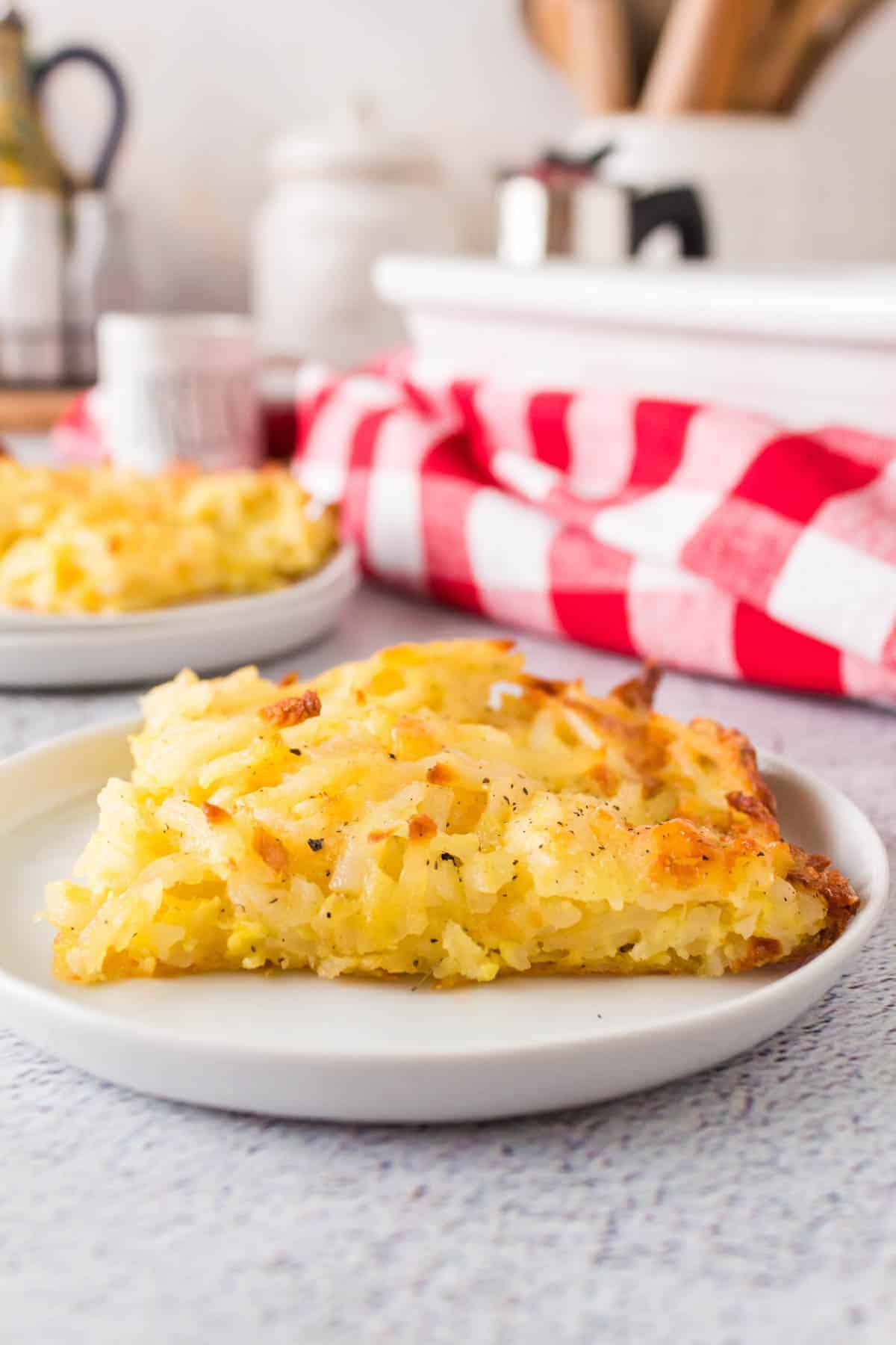 piece of the baked casserole on a white plate with a red and white checked napkin on a countertop with utensils in the background