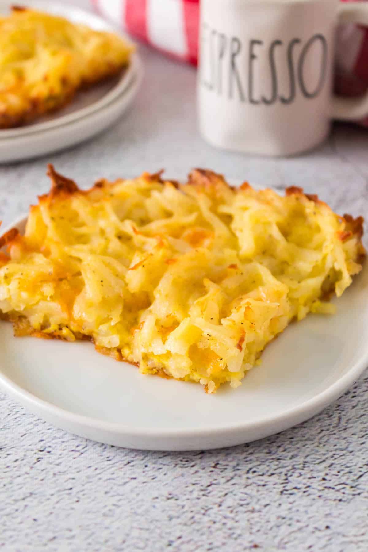 square of cheesy hashbrown casserole on a white plate in front of a coffee mug