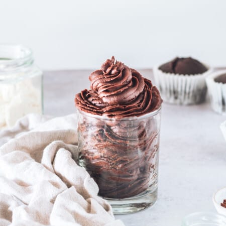 Homemade Chocolate Frosting Piped Into a Drinking Glass Beside Three Chocolate Cupcakes