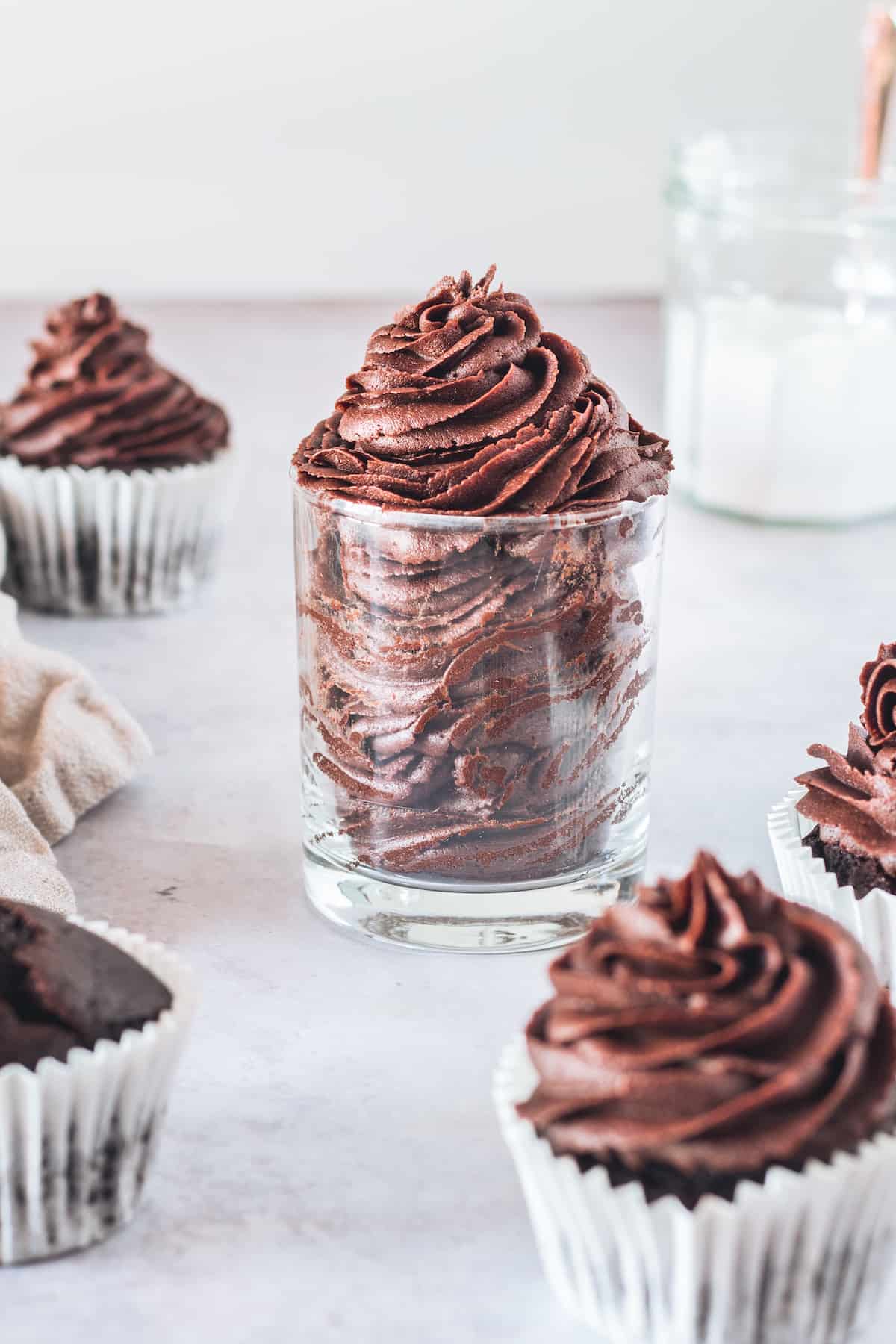 A Glass Filled with Homemade Chocolate Frosting on a Countertop with Frosted Cupcakes