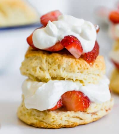 A strawberry shortcake with strawberries and whipped cream in the middle of two biscuit halves and on top with another in the background.