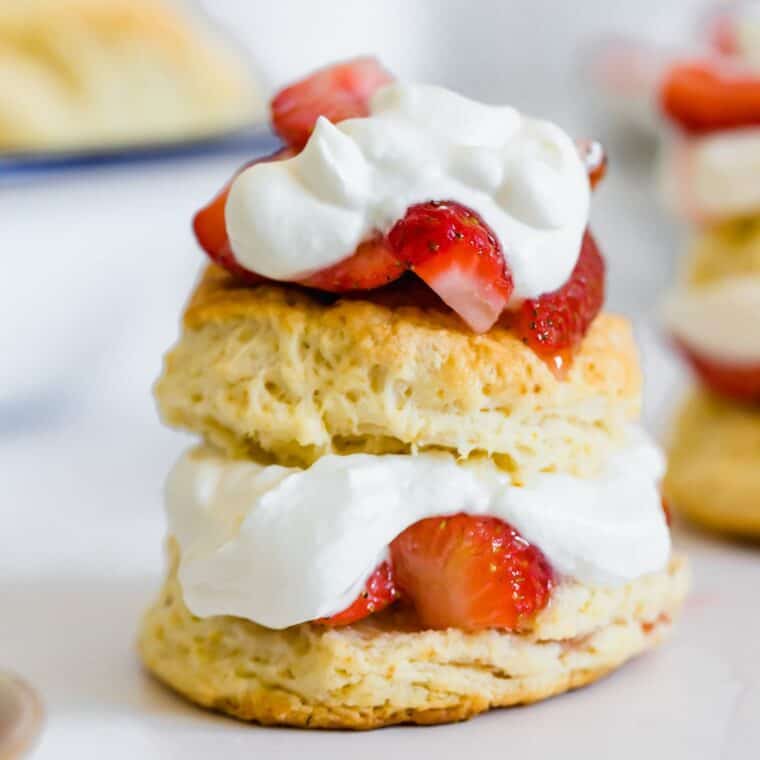 A strawberry shortcake with strawberries and whipped cream in the middle of two biscuit halves and on top with another in the background.