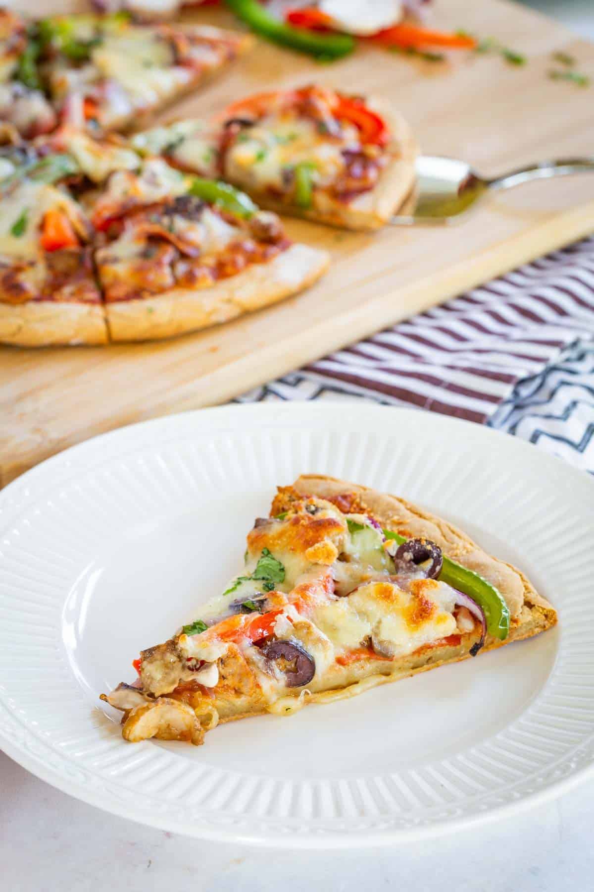 A Slice of Supreme Pizza on a White Plate with the Rest of the Pizza in the Background