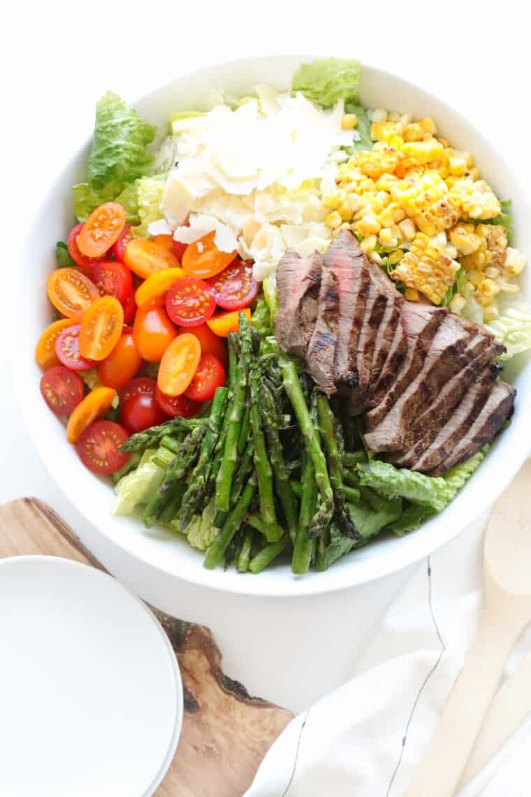 white serving bowl of grilled steak salad over romaine lettuce with tomatoes, parmesan and grilled asparagus and corn